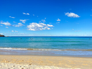 Fototapeta premium Surfers practicing catching a wave in the turquoise-blue ocean. Surf lesson. A day at the beach on a sunny day. Beach shore with sky and clouds.