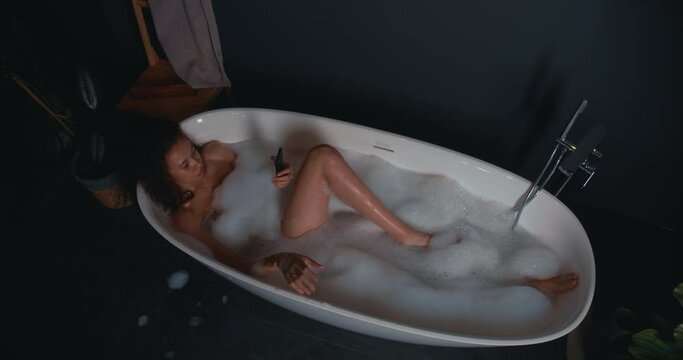 Relaxation concept. Beautiful young African American woman resting in modern bubble bath using smart phone shopping app.