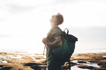 Carefree male traveler admiring view of sky near ocean shore with backpack - Powered by Adobe