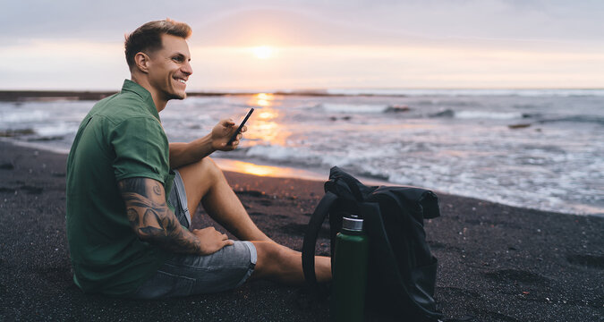 Smiling man in green shirt sitting on beach with smartphone in hand