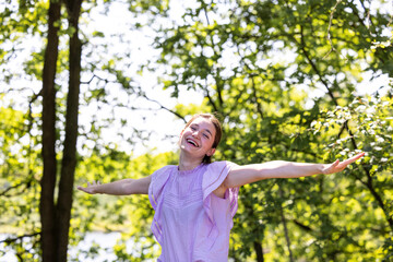 This photo depicts a woman in a lilac-colored top with her arms outstretched, face turned upwards, and eyes closed in a moment of pure bliss, as if embracing the warmth and beauty of the sunlit park