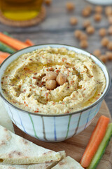 Traditional hummus made with chickpeas, olive oil, and garlic in a bowl with vegetable sticks and pita bread on wooden table. Vegan protein plant-based dip. 