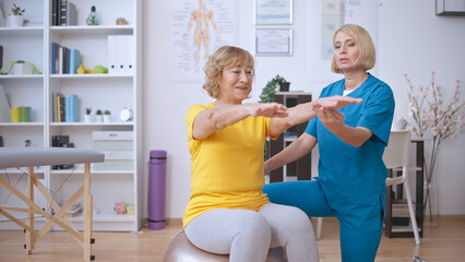 A female nurse assists a senior patient with exercises on a fitball in a rehabilitation center