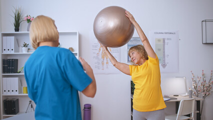 A positive mature woman leans right, holding a fitball in her hands above her head, rehabilitation...