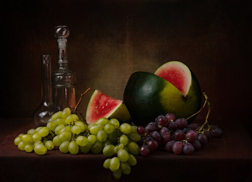 still life with a cut red watermelon with different varieties of green and black grapes and a couple of decorative bottles on the table on a dark background photo