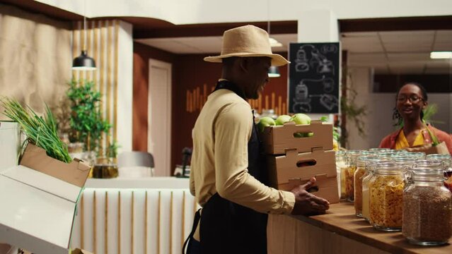 Local vendor placing crates with freshly harvested fruits on grocery store display, recommending organic produce to regular customer. African american farmer restocking all merchandise. Camera B.