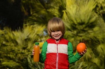 Cheerful child drinks orange juice. A boy in the garden, among the palm trees, holds an orange and a plastic bottle of juice. Healthy juices, vitamins for children, healthy eating