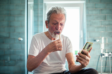 Senior man brushing teeth and using smartphone in bathroom