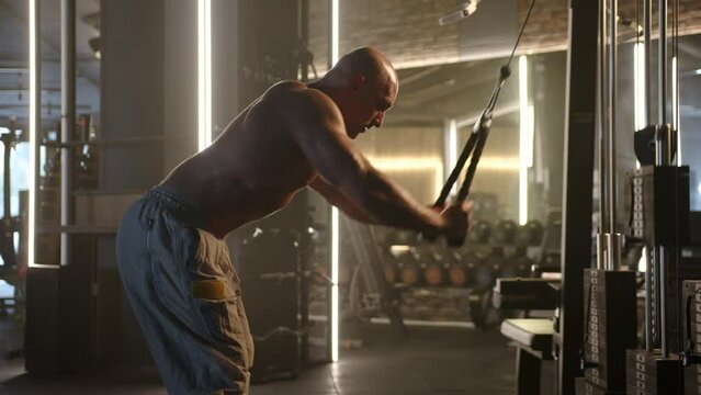 A bald man at the gym is fully concentrated on performing tricep pushdowns using a cable machine, targeting his upper arm muscles for a robust workout session. Camera 8K RAW. 