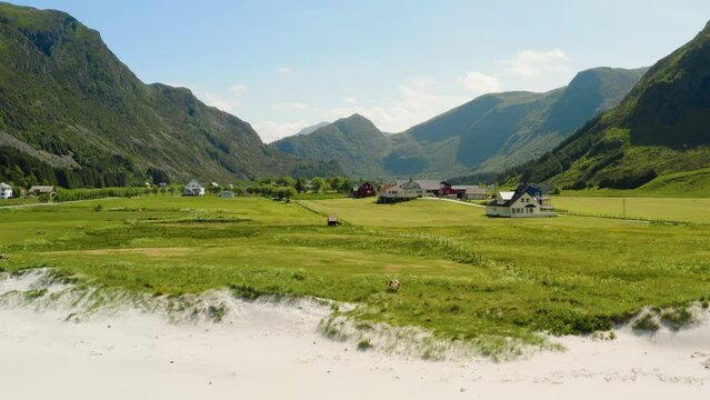 Cinematic aerial establishing shot, low drone flight pull in over white beach shore line towards small Refvik village green farm mountain landscape Summer vacation Refviksanden Norwegian Coast M&aring;l&oslash;y 