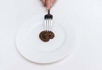 A hand with a fork is touching artificial feces lying on a white plate. The concept of an inept...