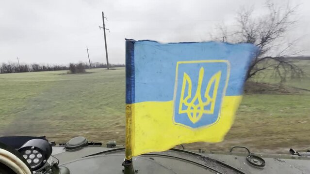 View from infantry fighting vehicle turret moving along a dirt road across field with fluttering Ukrainian flag on its.