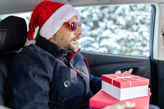 Fashionable man with santa hat and red sunglasses holding christmas presents while sitting on rear car seat. Winter scene in the background.