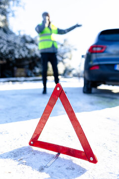 Winter season on the road car breakdown. Warning triangle on snowy road and man in safety vest using mobile phone in the background.
