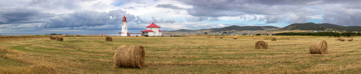 Wide panorama of the Anse a la Cabane lighthouse, the oldest working lighthouse on the Magdalen Islands, Gulf of Sait Lawrence, Canada. Rural setting with hay rolls in the foreground.