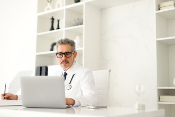 Mature, grey-haired doctor deeply engrossed in his work on a laptop, possibly consulting a medical database or researching for patient treatment, taking notes with serious expression reflecting