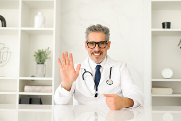 A genial mature doctor in a white coat waves hello, inviting a sense of comfort and accessibility,...