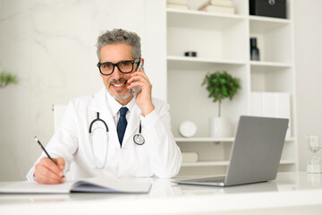 A seasoned doctor with grey hair is jotting down notes while attentively listening on a phone call, demonstrating multitasking and diligent patient care. His modern office reflects a professionalism