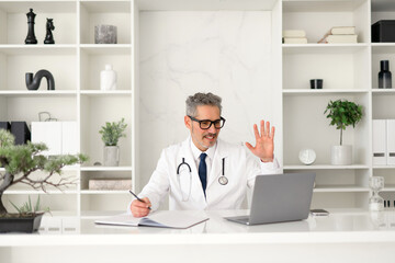 A grey-haired doctor waves in greeting, welcoming a patient to a virtual consultation, projecting a...