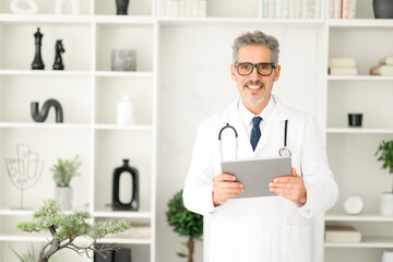 A mature, grey-haired doctor in a crisp white lab coat holds a digital tablet, poised to access...