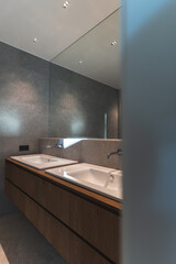Two spacious white bathroom sinks in front of a mirror with gray wall and wooden furniture