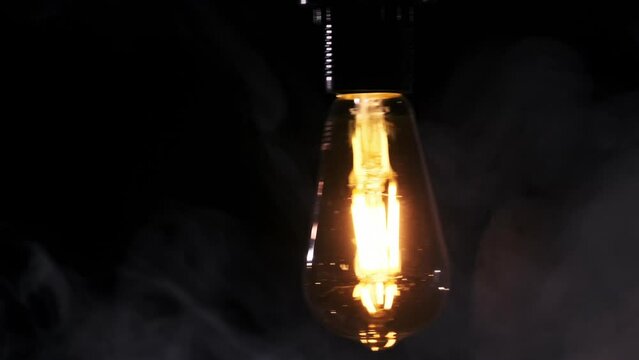 Transparent classic bulb hanging on wire and flickering on black background with smoke. Copy Space. Modern incandescent LED lamp glowing with warm filament. Tungsten on and off in dark. Vintage light.
