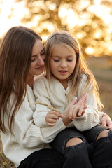 Fototapeta premium Family day, mother's day. Happy smiling young mom and adorable child daughter soft hugging, kissing and spending time together at autumn on sunset. Idyllic family having fun outdoors on fall holiday.