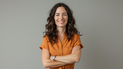 a cheerful young woman with long wavy hair, wearing an orange blouse, standing with her arms crossed against a neutral gray background