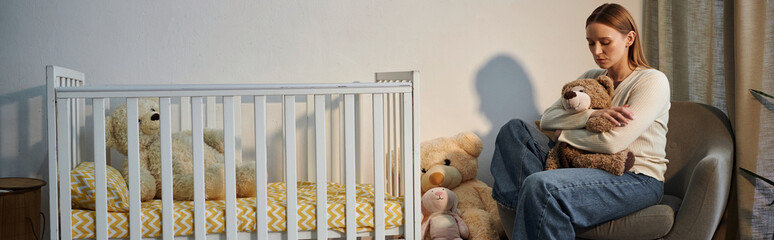depressed young woman with soft toy sitting in armchair near crib in dark nursery room, banner
