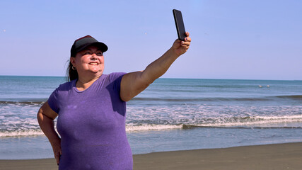 Portrait of an older, thick-set woman taking a selfie on the beach with the vastness of the sea behind her