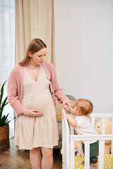 happy pregnant woman holding hand of little son standing in crib in cozy nursery room, motherhood