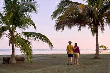 A couple standing on the beach facing the sea, both with beach attire; he wears a 'vueltiao' hat and she wears a visor. They have their backs to us, positioned between two palm trees