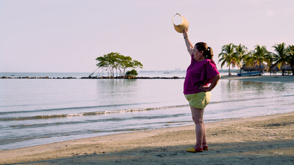 An older woman dressed in yellow shorts and a purple blouse standing sideways on the beach facing the sea, waving to someone in the distance with her raised hand holding her visor