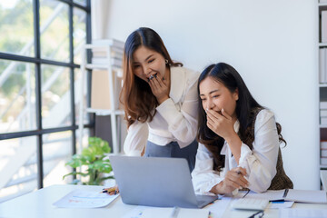 Two excited asian businesswomen celebrating a victory or good news in a modern office environment with a laptop and documents.