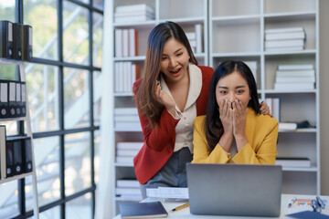 Two excited asian businesswomen celebrating a victory or good news in a modern office environment with a laptop and documents.