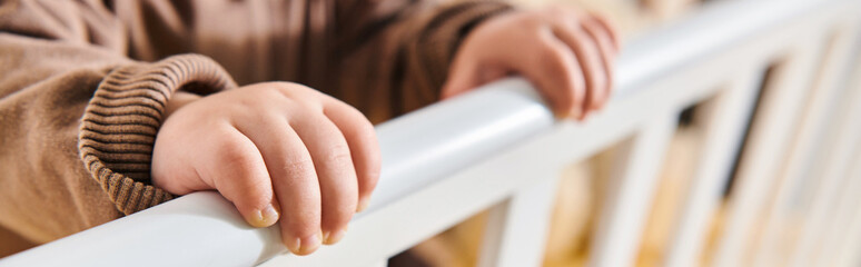 childhood, tiny hands of little kid standing in crib in nursery room at home, horizontal banner