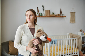 depressed young woman with soft toy looking away in nursery room at home, heartbroken moment