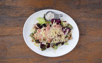 Top view of a Caesar salad with lettuce, parmesan cheese, bread croutons, salmon, bacon and Caesar dressing, in a white plate on the wooden table.