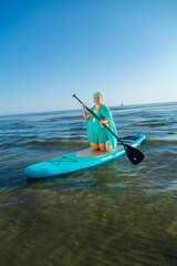 A woman in a turquoise swimsuit with a skirt and a scarf on her head on a SUP board near the seashore in the morning.