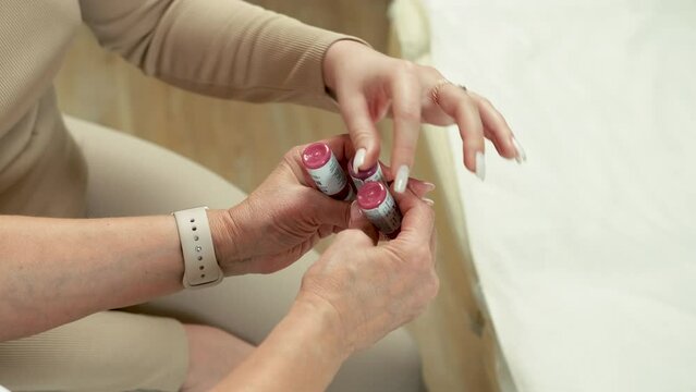 A client in a beige suit sits and chooses a pink pigment in a jar for permanent lip makeup in a beauty salon. The hands of a specialist with a watch on his wrist hold containers with pigment.
