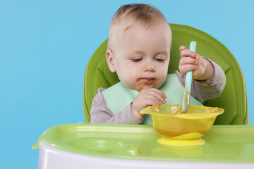 Cute little baby eating healthy food in high chair on light blue background