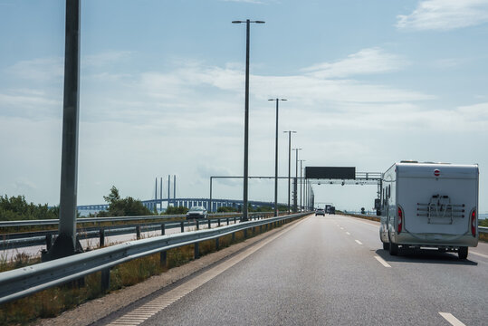 Traveling on a multilane highway, a white caravan is seen in the right lane under a partly cloudy sky. In the distance, the Oresund Bridge's pylons connect Copenhagen.