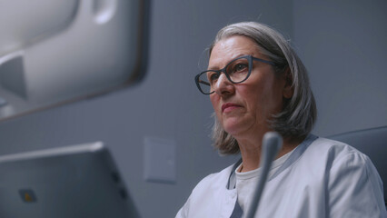 Mature female doctor looks at monitor of ultrasound machine while doing sonography diagnostics. Medical specialist performs check up or examination in modern clinic using advanced equipment. Portrait. - obrazy, fototapety, plakaty