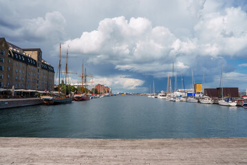 A serene harbor with a wooden dock leads to sailboats and motorboats in calm waters, under a dramatic, cloudy sky by historic buildings, likely in Copenhagen.