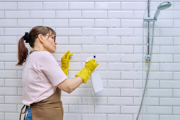 Middle-aged woman in an apron cleaning tiles in shower in bathroom