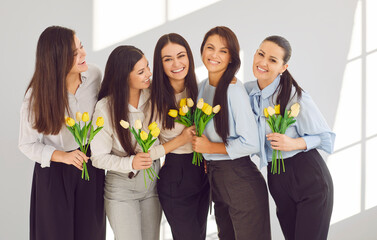 Group of happy business women in office celebrating International Women's Day together. Portrait of stylish and beautiful businesswomen with yellow tulips hugging and smiling in front of camera.