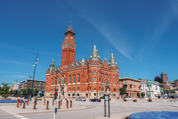 A sunlit red brick building with Romanesque and Gothic Revival elements, featuring a tower and arched windows, stands in a quiet plaza in Helsingborg.