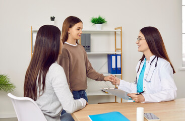 Fototapeta premium Child health. Caring female doctor takes hand of excited teenage girl who is worried before medical examination. Friendly doctor consults child who came with his mother for appointment at clinic.