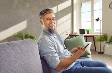 Man using smartphone. Happy handsome bearded senior man sitting on comfortable sofa in living room,...