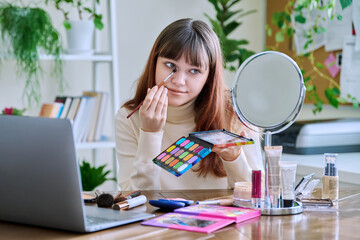 Young female showing cosmetics to laptop webcam, sitting at table at home